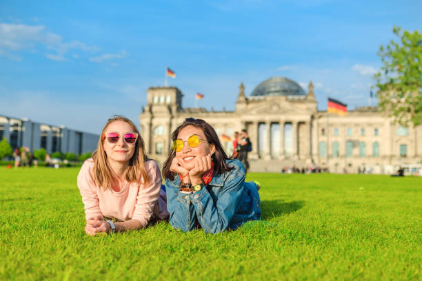 サングラスをかけた2人の若い幸せな女の子が芝生に横たわって、ベルリンの連邦議会ビルの前で楽しみを持っています。留学とドイツ旅行のコンセプト - the reichstag ストックフォトと画像
