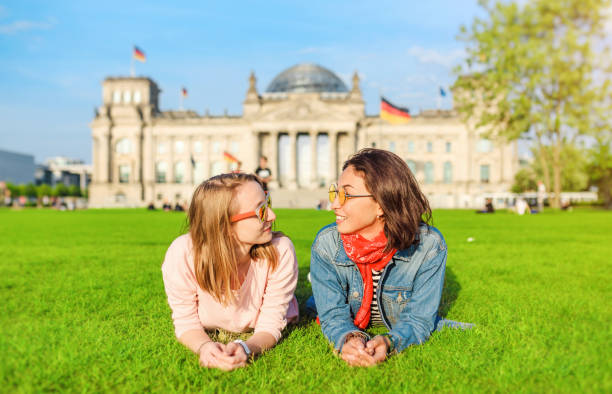 deux jeunes filles heureuses portant des lunettes de soleil se trouvant sur une pelouse et amusez-vous devant le bâtiment du bundestag à berlin. étudier à l’étranger et voyager en allemagne concept - glasses grass women lying on front photos et images de collection