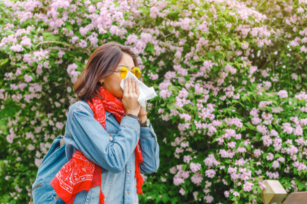 Allergy concept. Young asian woman sneezes and blowing her nose with a handkerchief and suffering in the spring among flowering and blooming trees. stock photo