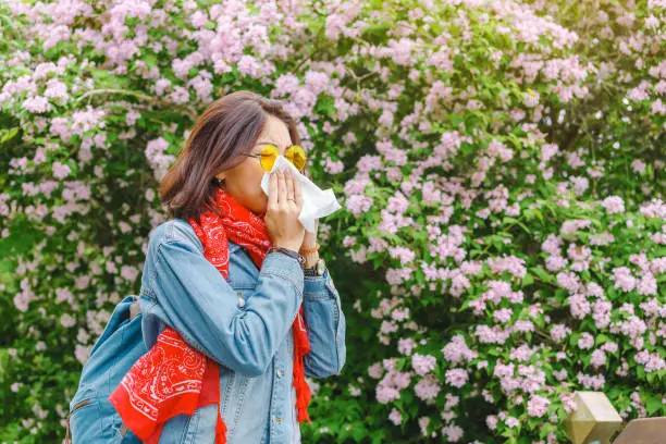 Photo of Allergy concept. Young asian woman sneezes and blowing her nose with a handkerchief and suffering in the spring among flowering and blooming trees.