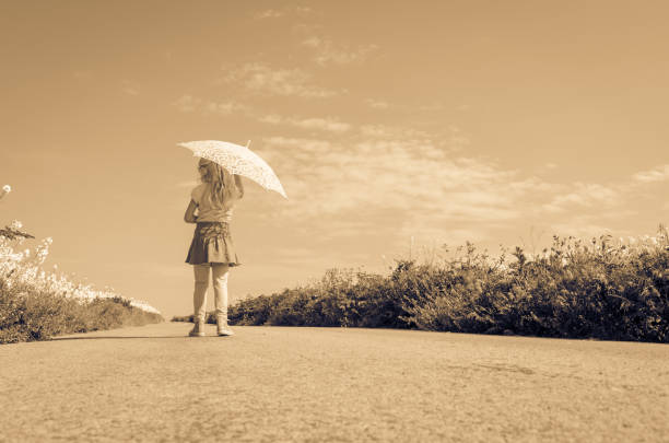teenage girl with umbrella walking among yellow rapeseed fields in empty rural path sepia tone