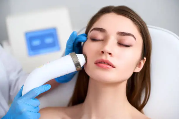 Beautician doctor doing rf-lifting procedure for flawless woman face laying on medical chair in a beauty salon. Hardware cosmetology. Patient receiving electric facial massage. Skin rejuvenation and wrinkle smoothing.