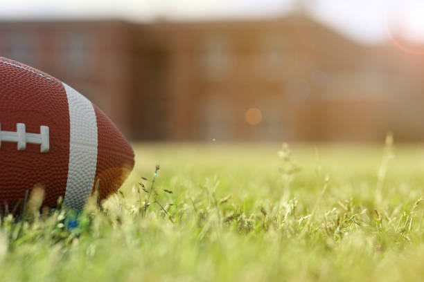Football on grass stadium on college or high school campus. School building in background. No people. Daytime.