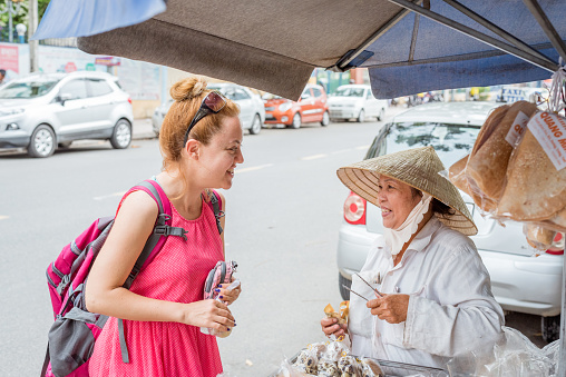 Nha Trang, Vietnam - May 5, 2018: a solo European traveler and a street dessert vendor, a Vietnamese lady in an Asian conical hat, happily look at each other after completing the shopping interaction.