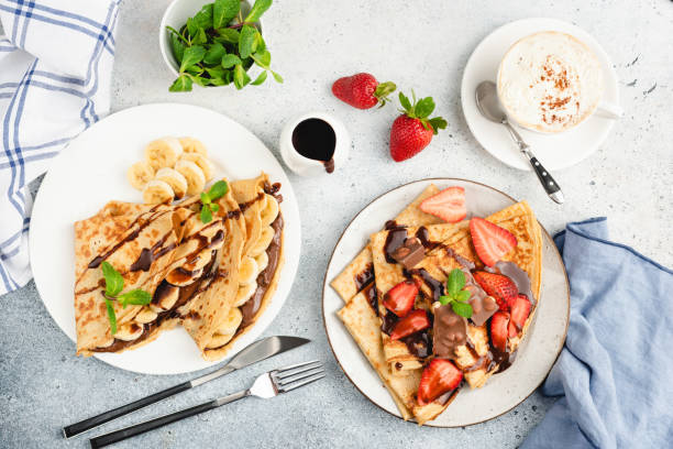 Crepes or blini with fruits, cup of coffee Crepes or blini with banana, strawberry and chocolate, cup of cappuccino coffee and decoration of mint leaf and chocolate sauce. Top view, flat lay composition crêpe pancake stock pictures, royalty-free photos & images