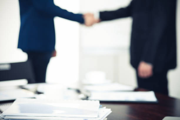 istock Documents and laptop on the table. Business people shaking hands on the background, silhouettes 1145770535