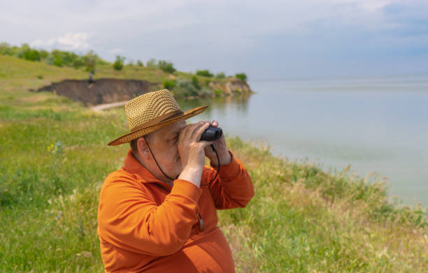 ritratto di uomo anziano con cappello di paglia che guarda nel binocolo mentre è seduto sul fiume dnipro durante la stagione primaverile - binoculars watching optical instrument closed foto e immagini stock