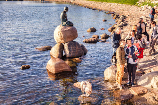 23 September 2018: Copenhagen, Denmark - Asian tourists taking selfies with a selfie stick at the  Little Mermaid, the famous bronze statue by Edvard Eriksen, which was based on the fairy tale of the same name by Danish author Hans Christian Andersen. It was erected in 1913.
