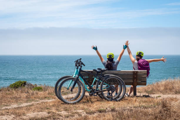 велосипедист сестры сидят на скамейке с поднятыми руками, просмотр океана - half moon bay стоковые фото и изображения