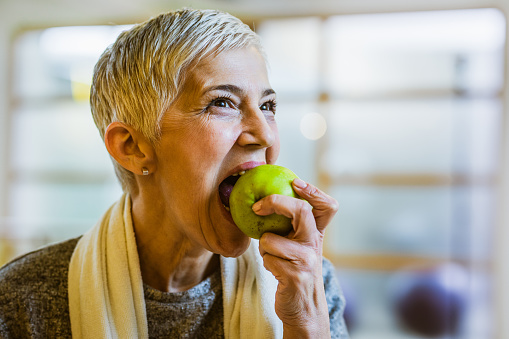 Smiling senior woman biting an apple after sports training in a health club.