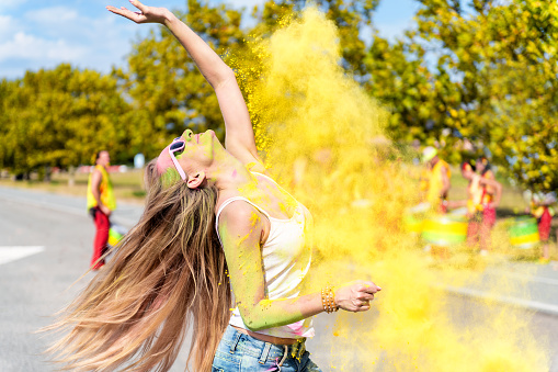 Woman loving the color festival, feeling free and dancing. Colored yellow powder exploding and falling on her. Young candid girl with face paint wearing pink sunglasses and enjoying the event. Drummer Indian musicians in the background.
