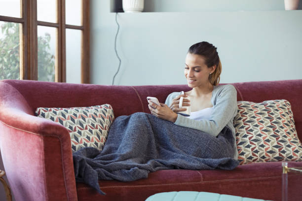 istock Pretty young woman using her mobile phone while drinking coffee on sofa at home. 1145837553