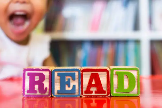 Photo of Alphabet blocks spelling the word read in front of a bookshelf and an excited child in the background