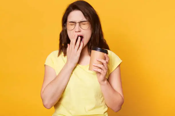 Portrait of beautiful charismatic young female holding papercup with coffee, covering mouth while yawning, closing eyes. Brunette European model wears casual yellow t shirt and stylish spectacles.