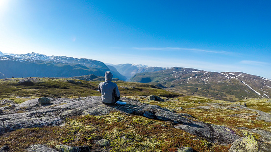A young man wearing a hoodie sit at the ground and enjoys the endless mountain view of Eidfjord. He is relaxed. In the back taller mountains are covered partially with snow. Clear and sunny day.