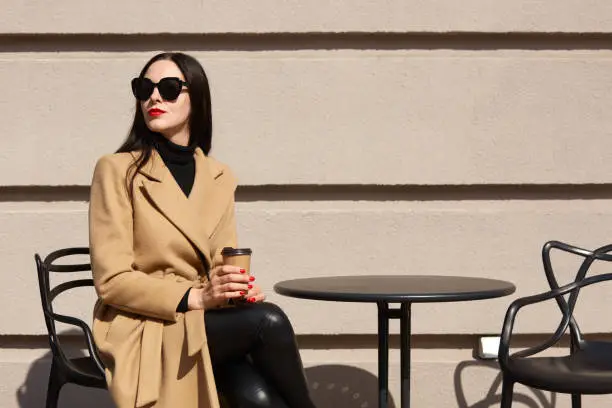 Charming stylish model with red lipstick wearing big trendy sunglasses, beige coat, black sweater and trousers, holding papercup of strong coffee, sitting outdoors over light wall, enjoying sunlight.