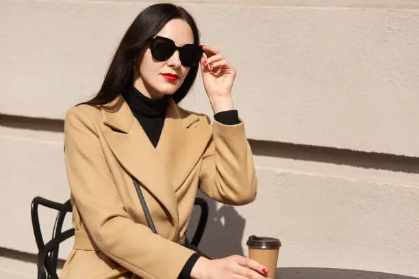 Black haired European magnetic female holding brown papercup in one hand, touching black sunglasses, looking aside. Beautiful energetic model wearing stylish clothes, having nice rest outside.
