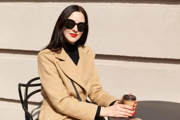 Smiling fashionable businesswoman with bright red lips sitting in bright wooden chair, holding papercup of strong coffee, wearing beige coat, black sweater and accessories. Street fashion concept.