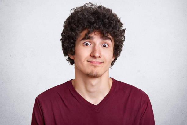 fermez le portrait du modèle mâle beau avec les cheveux nets, posant avec l’expression faciale de malentendu, porte le t-shirt marron occasionnel, se dresse contre le mur en béton blanc. concept d’émotions de personnes. - male maroon head and shoulders close up photos et images de collection