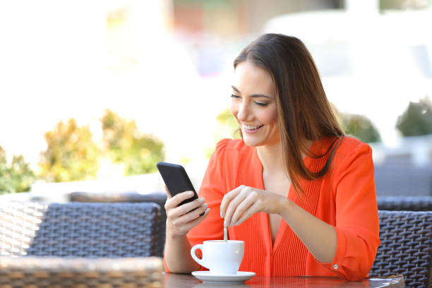 Happy woman using phone stirring coffee in a bar stock photo