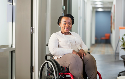 A mid adult African-American businesswoman in a wheelchair in an office hallway, smiling and looking at the camera. Her disability is the result of having spina bifida.