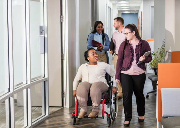 Business people going to meeting, woman in wheelchair A multi-ethnic group of four business people walking down an office corridor, on their way to a meeting, carrying, notebooks and files. The focus is on the two women in front, an African-American woman in her 30s in a wheelchair with spina bifida, and her young Hispanic coworker, in her 20s. They are chatting. persons with disabilities photos stock pictures, royalty-free photos & images