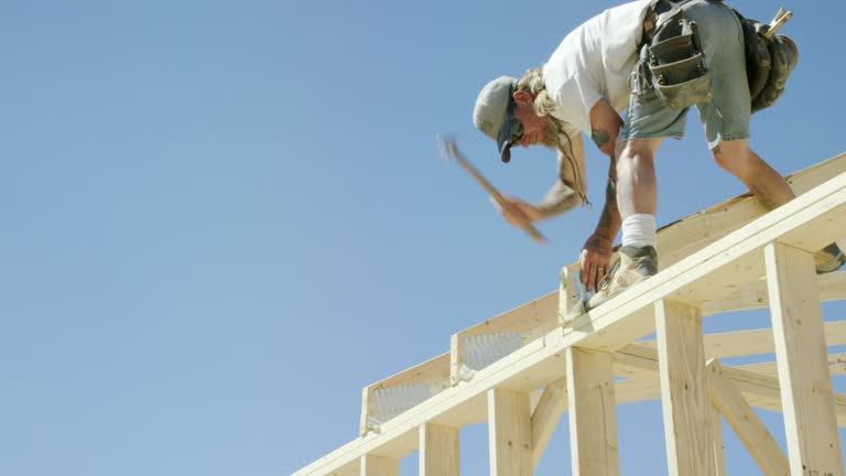 A Caucasian Male Construction Worker in His Forties with Tattoos Secures a Framed Wooden Roof Truss by Hammering a Nail to the Structure While Framing a House on a Clear, Sunny Day