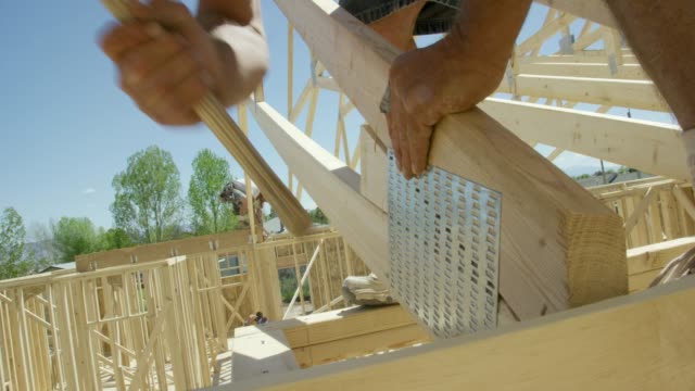 Two Caucasian Male Construction Workers in Their Forties Attach a Framed Wooden Roof Truss to a Structure Using Hammers and Nails While Framing a House and Smoking on a Clear, Sunny Day
