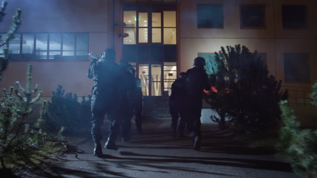 Masked Team of Armed SWAT Police Officers Run in Formation Forwards an Office Building. Soldiers with Rifles and Flashlights Run on a Street Filled with Smoke.