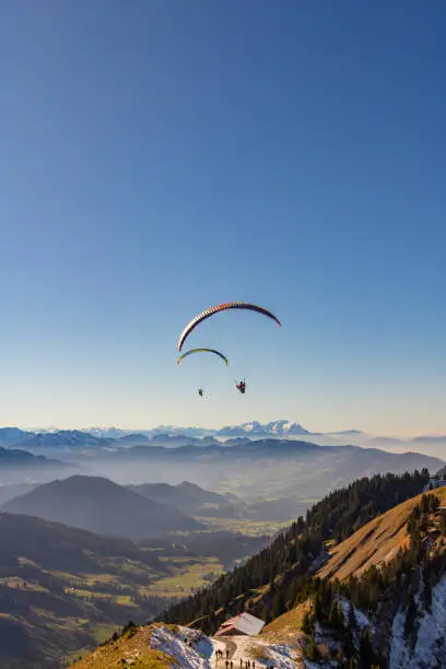 Paragliders flying straight to a valley over a hut with people around in Oberstaufen, Germany.