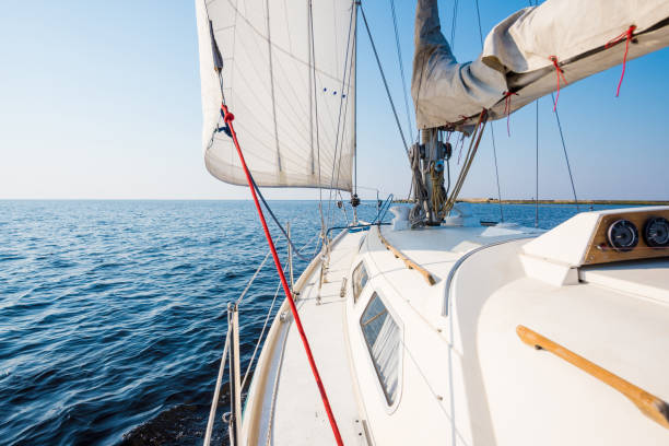 White yacht sails on a clear sunny day. A view from the deck to the bow and sails, close-up. Dolphins and lighthouse in the background. Baltic sea, Latvia