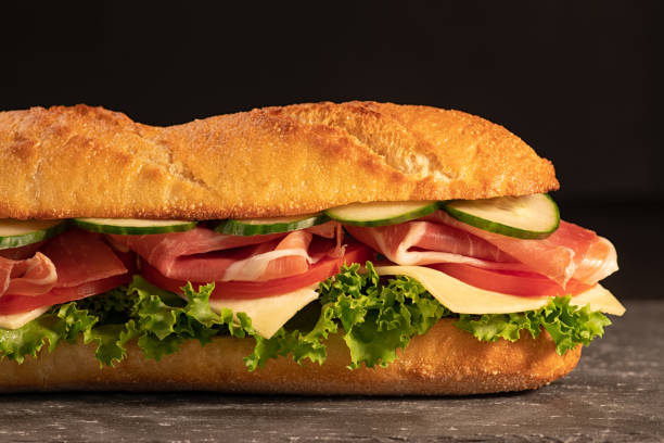 istock Huge fresh crispy baguette sandwich with meat, prosciutto, cheese, lettuce salad and vegetables. Close up. Black background. Space for text. 1145893733