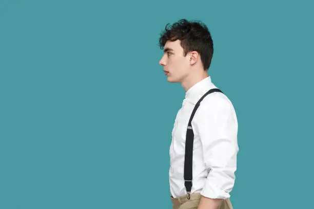 Profile side view portrait of serious handsome hipster curly young businessman in classic casual white shirt and suspender standing and looking straight. indoor studio shot isolated on blue background
