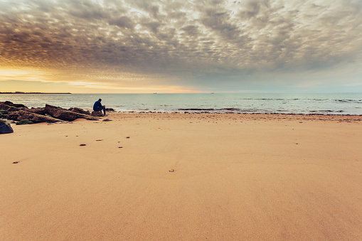 Caucasian man alone sitting on the shore of the sea, contemplating the sunset. Thoughts and loneliness of a man on the beach