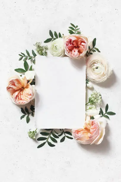 Photo of Feminine wedding, birthday mock-up scene. Blank paper greeting card. Floral frame of blush pink English roses, ranunculus flowers and lentisk leaves. Concrete table background. Flat lay, top view.