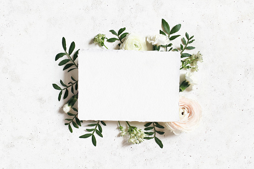Feminine wedding, birthday mock-up. Blank paper greeting card. Floral frame of white, pink ranunculus, carnation and astrantia flowers, lentisk leaves, concrete table background. Flat lay, top view.