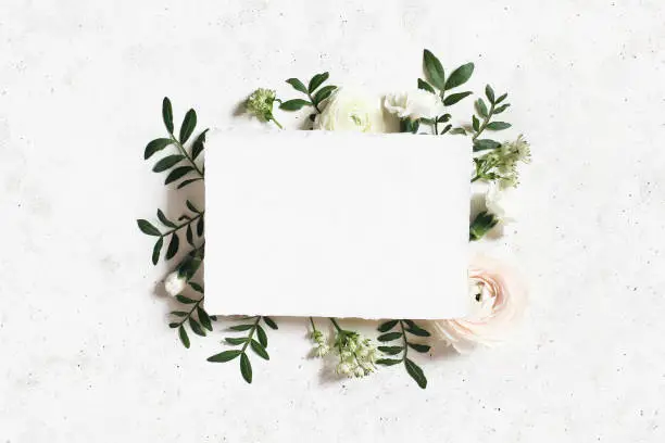 Photo of Feminine wedding, birthday mock-up. Blank paper greeting card. Floral frame of white, pink ranunculus, carnation and astrantia flowers, lentisk leaves. Concrete table background. Flat lay, top view.