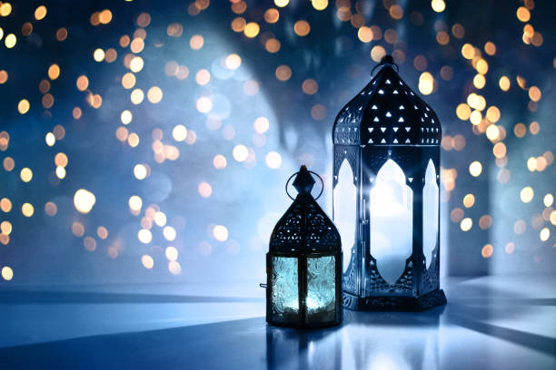 Couple of glowing Moroccan ornamental lanterns on the table. Greeting card, invitation for Muslim holy month Ramadan Kareem. Festive blue night background with glittering golden bokeh lights. stock photo