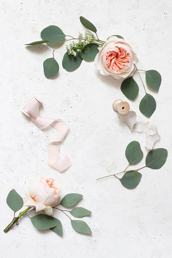 Feminine wedding, birthday still life scene. Silk ribbons, eucalyptus leaves and blush pink English roses flowers. Concrete table background. Flat lay, top view,vertical, empty copy space.