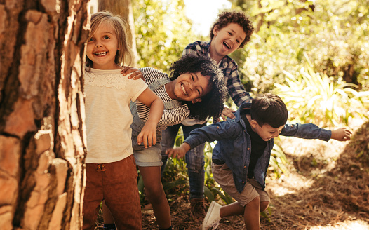 Cute smiling kids peeking out from behind the tree in the park. Group of children enjoying playing hide and seek in a forest.