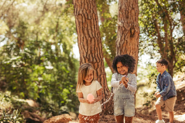 Kids playing with water balloon in park Girls closes eyes while bursting water balloons. Water droplets fly through the air and the kids have fun in park. water balloon stock pictures, royalty-free photos & images