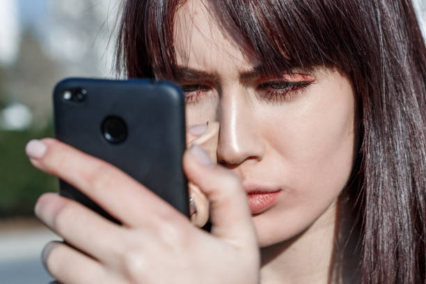 istock A young and beautiful woman adjusts her makeup as she uses her smartphone camera as a mirror. She is standing outdoors during the day. 1145917349