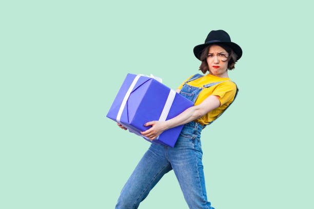 confused strong young girl in fashionable hipster wear in denim overalls and black hat standing and trying to hold giant big heavy gift box with fragile face. - violence hat toughness blue imagens e fotografias de stock