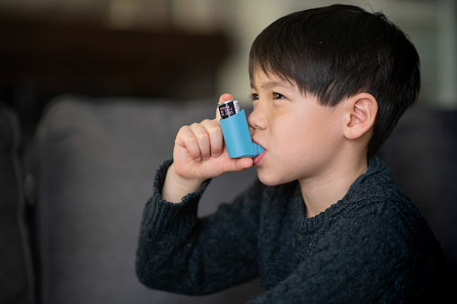 A little boy is sitting in a living room. He is using puffer because he suffers from asthma.