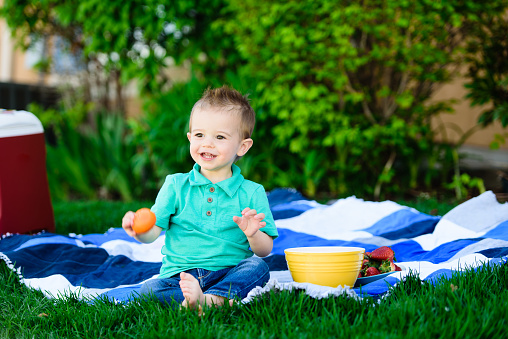 Preschool age boy sitting on blue and white picnic blanket, holding a clementine, smiling and looking away.