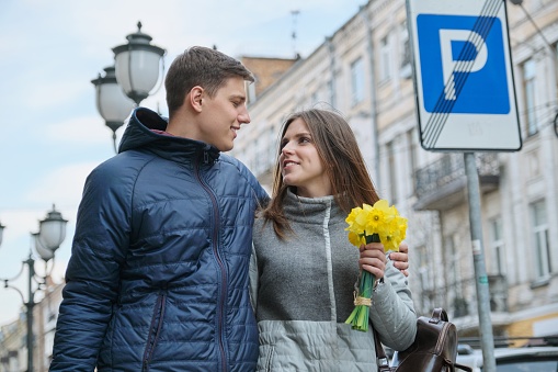 Spring outdoor portrait of young couple walking with bouquet of yellow spring flowers daffodils, happy youth, city street background.