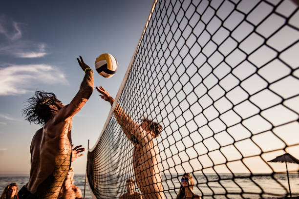 Young man blocking his friend while playing beach volleyball in summer day. Group of friends having fun while playing volleyball on the beach. One man is trying to block his friend. male volleyball player stock pictures, royalty-free photos & images