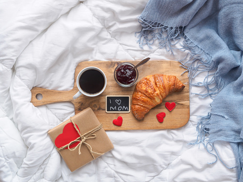 Breakfast with coffee, croissant and jam on a cutting board on a white coverlet, top view, birthday present, paper heart