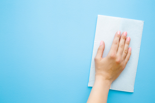 Woman's hand wiping pastel blue desk with white paper napkin. General or regular cleanup. Close up. Empty place for text or logo. Top view.