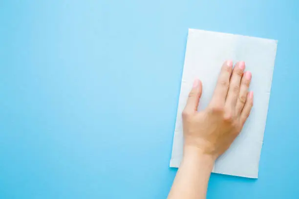 Photo of Woman's hand wiping pastel blue desk with white paper napkin. General or regular cleanup. Close up. Empty place for text or logo. Top view.